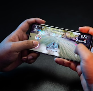 Hands holding a smartphone with a racing game on the screen, featuring a car speeding along a track with visible game controls and speed indicators.